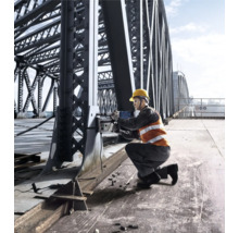 Un muncitor în construcții, cu cască de protecție și vestă de siguranță, operează o mașină de găurit magnetică pe un pod de oțel.