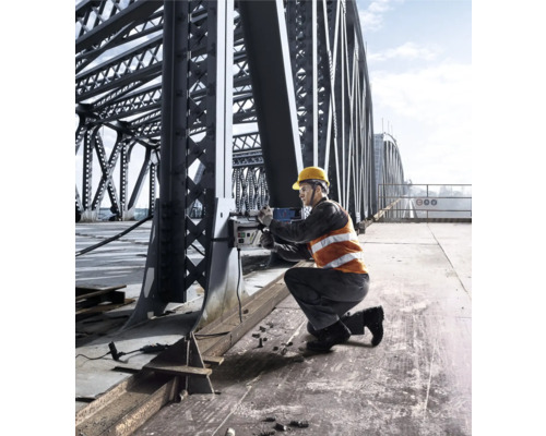 Un muncitor în construcții, cu cască de protecție și vestă de siguranță, operează o mașină de găurit magnetică pe un pod de oțel.