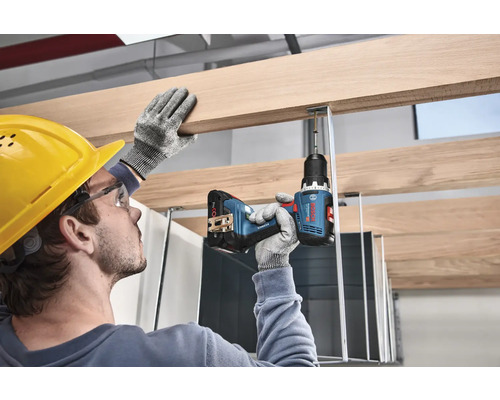 Bărbat cu cască de protecție și ochelari de protecție lucrează cu o șurubelniță cu acumulator la un raft de lemn. Logo Bosch.