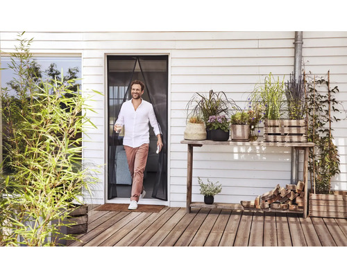 Un bărbat trece printr-o perdea magnetică împotriva insectelor la o ușă de terasă pe o terasă din lemn cu diverse plante la ghiveci.