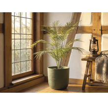 Palmier artificial într-un ghiveci verde texturat într-o cameră rustică luminoasă, cu grinzi de lemn, o fereastră și un taburet cu felinar.