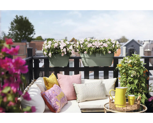 Jardiniere de balcon de culoare verde salvie cu flori roz deschis pe o balustradă neagră deasupra unei canapele deschise cu perne colorate.
