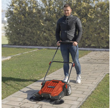 O persoană împinge o mașină de măturat Einhell pe o alee de grădină pavată, lângă un gazon și un gard viu.