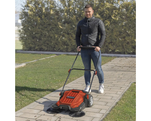 O persoană împinge o mașină de măturat Einhell pe o alee de grădină pavată, lângă un gazon și un gard viu.