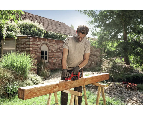 Un bărbat folosește o rindea electrică pe o grindă de lemn pe capre într-o grădină luminoasă. Poartă ochelari de protecție și mănuși.