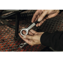 Mâini care țin un dispozitiv de tăiat piulițe din metal într-un atelier deasupra unui grătar metalic.