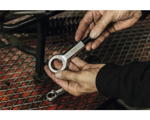 Mâini care țin un dispozitiv de tăiat piulițe din metal într-un atelier deasupra unui grătar metalic.