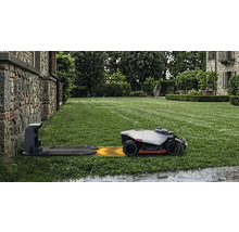 Robot de tuns iarba Mammotion pe gazon verde se apropie de stația de încărcare pe ploaie în fața unui zid de piatră.