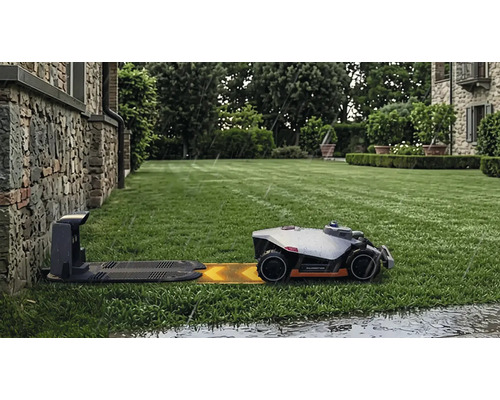 Robot de tuns iarba Mammotion pe gazon verde se apropie de stația de încărcare pe ploaie în fața unui zid de piatră.