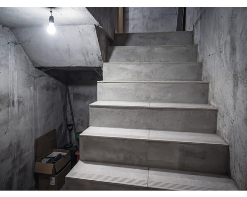 Scară din beton într-o pivniță cu pereți nefinisați, luminată de un singur bec.