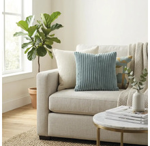 Sufragerie luminoasă cu canapea textilă, perne decorative din catifea raiată, pătură, măsuță rotundă și plantă de interior lângă fereastră.