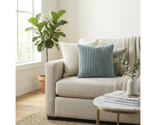 Sufragerie luminoasă cu canapea textilă, perne decorative din catifea raiată, pătură, măsuță rotundă și plantă de interior lângă fereastră.