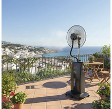 Ventilator mobil cu pulverizare pe o terasă însorită, cu vedere la mare și la un oraș de coastă, mobilier din lemn și plante în ghivece.