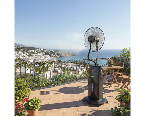 Ventilator mobil cu pulverizare pe o terasă însorită, cu vedere la mare și la un oraș de coastă, mobilier din lemn și plante în ghivece.