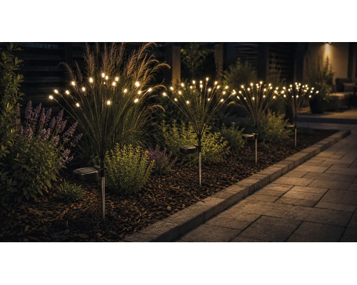 Lămpi solare de grădină cu design de artificii și becuri LED cu lumină albă caldă într-un strat de flori lângă o alee pavată.