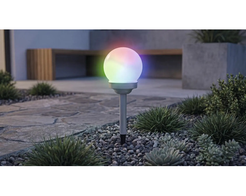 Lampă solară de grădină în formă de glob cu iluminare RGB multicoloră, pe un țăruș într-un strat de pietriș cu plante.
