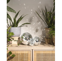 Trei globuri de oglinzi de dimensiuni diferite pe o bufetă din lemn și ratan, înconjurate de plante de apartament și reflexii de lumină pe perete.