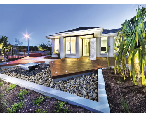 Casă modernă cu terasă din lemn iluminată, grădină de piatră cu element de apă și plante tropicale la amurg.