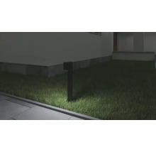 Lampă de grădină neagră modernă care luminează o zonă cu iarbă lângă o alee pavată și un perete de casă noaptea.