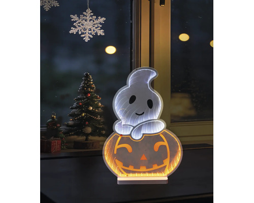 Decorațiune de Halloween cu LED fantomă pe dovleac cu efect de oglindă infinită în fața unei ferestre lângă un brad de Crăciun împodobit.