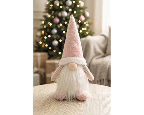 Pitic din material textil cu căciulă ascuțită și barbă pe o masă de lemn într-o cameră luminoasă, decorată festiv cu un brad de Crăciun.