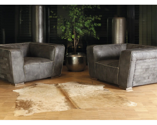Două fotolii din piele gri cu cusături decorative pe parchet, un covor din piele de vacă și o plantă verde într-un ghiveci metalic.