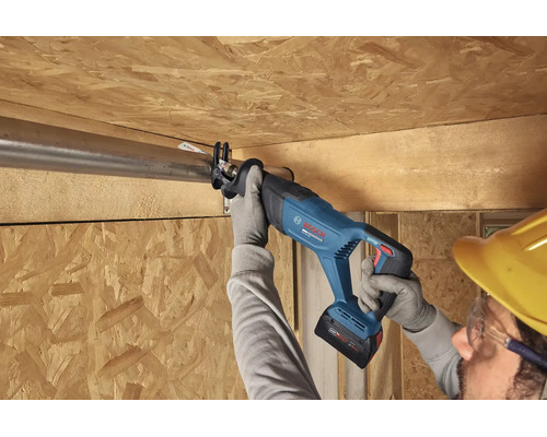 Meșter cu cască de protecție și mănuși taie o țeavă metalică cu un ferăstrău sabie cu acumulator Bosch la o construcție de tavan din lemn.