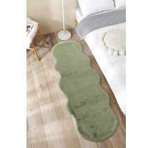 Traversă din blană artificială verde cu margini ondulate într-un dormitor luminos pe podea de lemn lângă un pat.