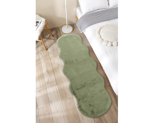 Traversă din blană artificială verde cu margini ondulate într-un dormitor luminos pe podea de lemn lângă un pat.