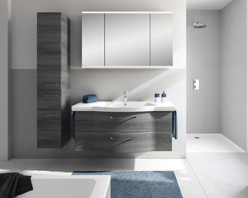 Set modern de mobilier de baie cu mască de chiuvetă închisă la culoare, lavoar alb, dulap cu oglindă cu trei uși și dulap înalt în baie luminoasă.