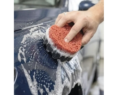 O mână spală o mașină albastru închis cu un burete auto stratificat și spumă de curățare.