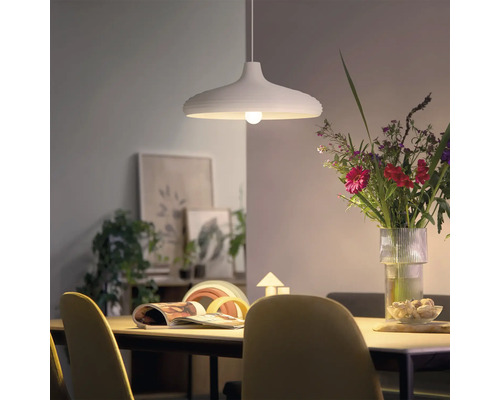 Lampă suspendată modernă deasupra unei mese de dining cu un buchet de flori și decorațiuni în fundal, într-o cameră luminoasă.