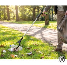 O persoană folosește un clește pentru gunoi într-un parc pentru a ridica o sticlă de plastic de pe sol și a o pune într-un sac de gunoi. (Image AI modified or generated)
