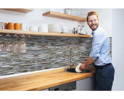 Tapet d-c-fix Ceramics Stone wall gri 67,5x400 cm Scenă de bucătărie cu panouri de perete din piatră, chiuvetă și bărbat care spală vase