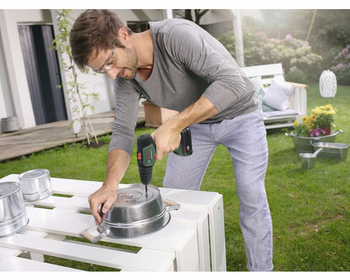 Bărbat găurind o găleată metalică în grădină cu o șurubelniță electrică.