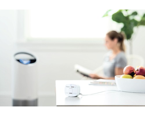 Scenă cu purificator de aer și senzor pe o masă, cu o femeie citind în fundal