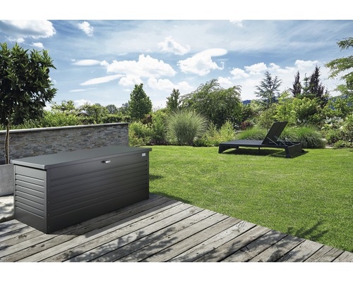 Scenă de grădină cu o cutie de depozitare metalică pe o terasă de lemn și un șezlong pe gazon