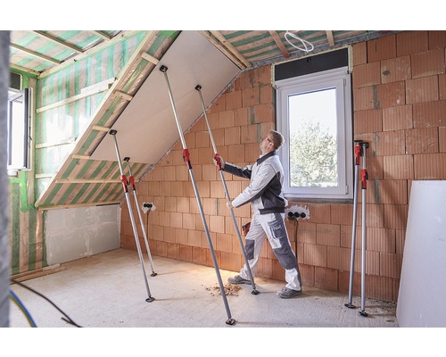 Un meșter montează o placă de gips-carton cu suporturi telescopice într-o mansardă în construcție.