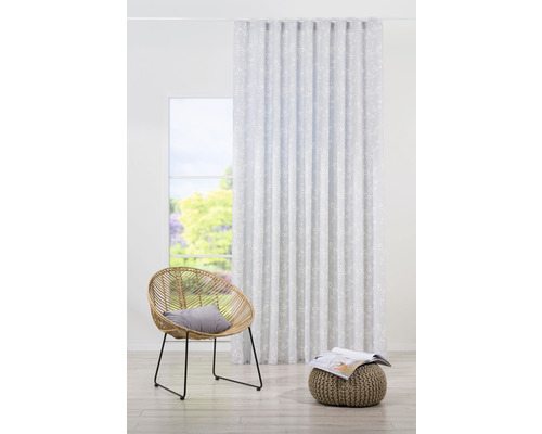 Scenă cu o perdea albă cu model floral, un scaun de ratan și un taburet tricotat într-o cameră luminoasă.