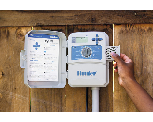 Controler de irigare Hunter X2 cu carcasă deschisă pe un perete de lemn, o mână operează controlerul