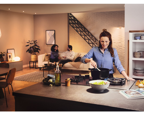 Bucătărie luminoasă, deschisă, cu o femeie care gătește la aragaz, în timp ce două persoane stau pe o canapea