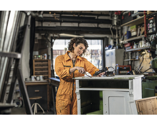 Femeie îndepărtând vopseaua cu un pistol cu aer cald de pe un dulap de lemn într-un atelier.