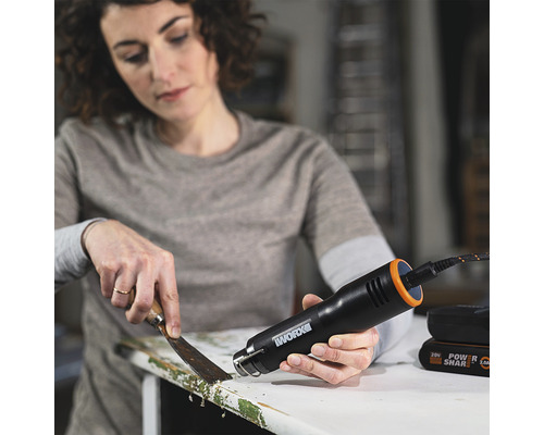 O femeie îndepărtează vopseaua de pe o piesă de mobilier cu un pistol cu aer cald și o spatulă.