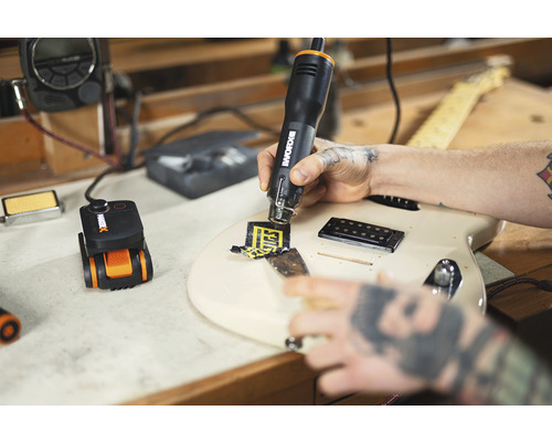 Un bărbat îndepărtează un autocolant de pe o chitară cu un pistol cu aer cald. Un instrument de încălzire de precizie se află pe un banc de lucru.