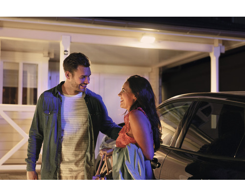 Un cuplu zâmbind în fața unei case și a unei mașini.