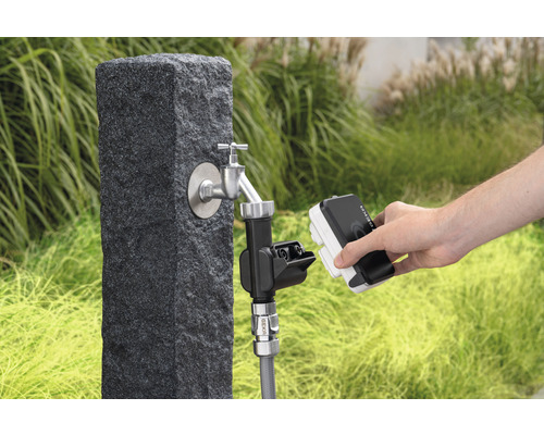 Scenă de grădină cu robinet pe un soclu de piatră și o persoană care conectează un dispozitiv de irigare