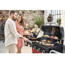 Persoane care fac grătar cu un grătar pe gaz pe o terasă cu iluminare și un zid de cărămidă în fundal.