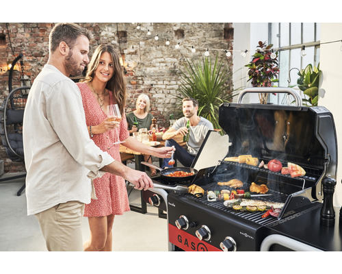 Persoane care fac grătar cu un grătar pe gaz pe o terasă cu iluminare și un zid de cărămidă în fundal.