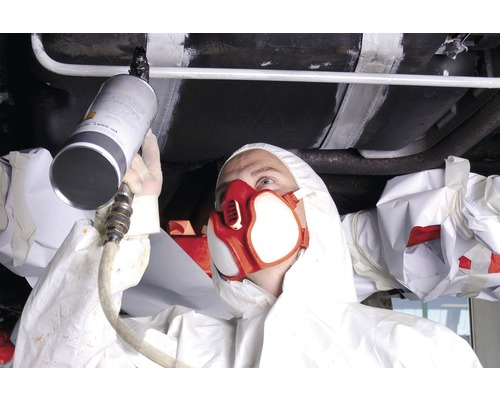 O persoană în echipament de protecție pulverizează un strat de protecție sub caroserie pe o mașină.