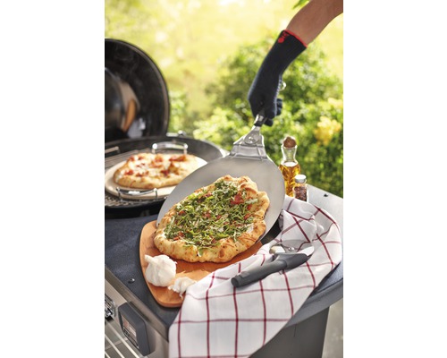 O persoană pregătește pizza în aer liber pe un grătar cu o paletă de pizza, un tocător, un prosop de bucătărie și usturoi.